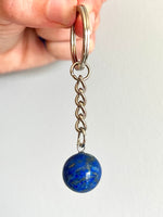 A close-up of a hand holding a keychain with a silver-toned split ring, short chain, and a round blue lapis lazuli stone pendant against a plain light background.