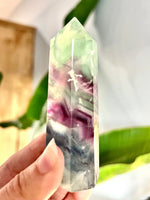 A hand holding a polished natural fluorite crystal point with marbled green, purple, white, and blue tones, set against a softly blurred background with green leaves.