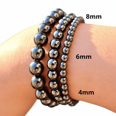 Three beaded stretch bracelets in different sizes (8mm, 6mm, and 4mm) worn on a wrist. The bracelets are made of polished black beads with a metallic sheen, reflecting the surrounding environment.