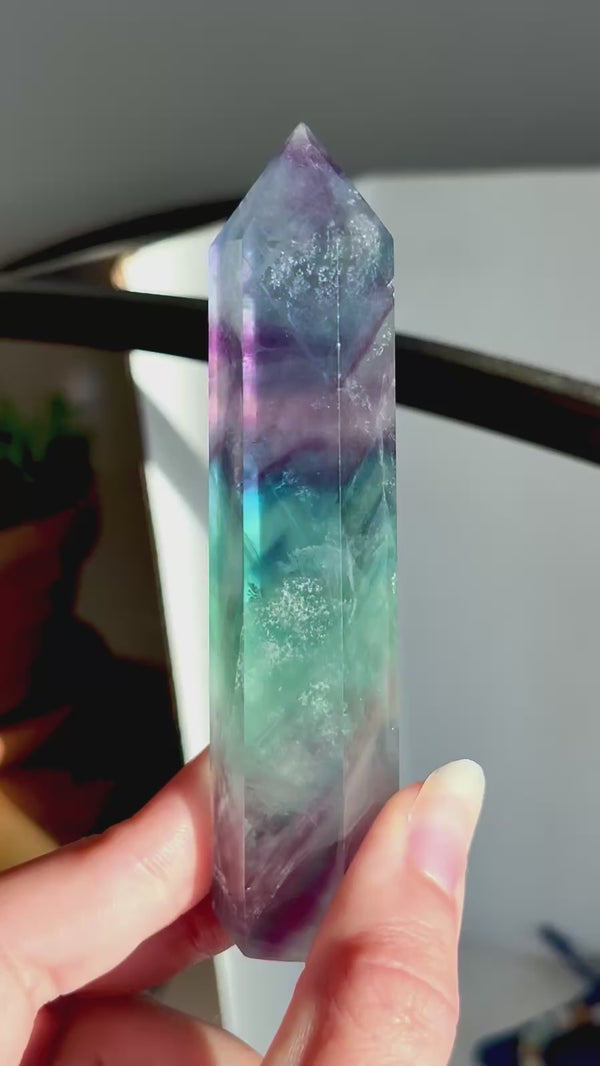A hand holding a polished natural fluorite crystal point with marbled green, purple, white, and blue tones, set against a softly blurred background with green leaves.