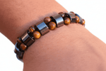 A close-up of a person's wrist wearing a beaded bracelet with brown tiger's eye beads, black spacer beads, and shiny silver metallic links.